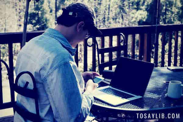 Молодой человек смотрит на смартфон, ноутбук, кафе на террасе