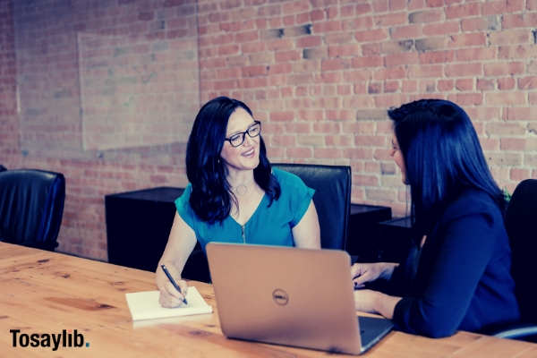 two woman sitting talking laptop notebook ballpen table chair