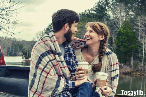 couple holding coffee covered with blanket sitting back truck