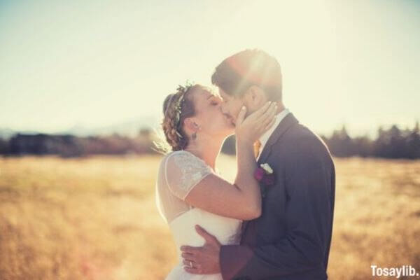 mulher vestindo um vestido de casamento beijos de um homem vestindo um terno cinza no meio de um campo em frente