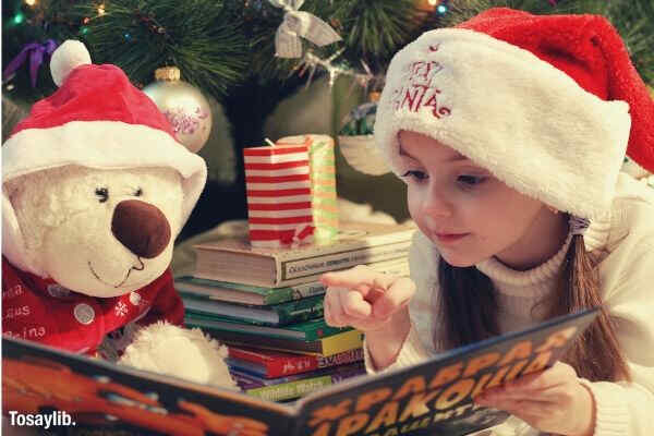 portrait of girl wearing christmas hat reading teady bear christmas tree