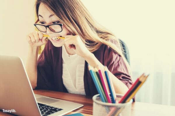 woman biting pencil sitting on chair looking laptop ballpens table