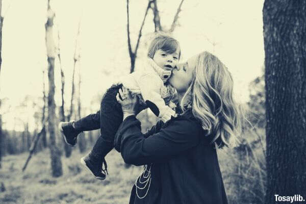 grayscale photo of woman kissing toddler on cheek standing beside tree
