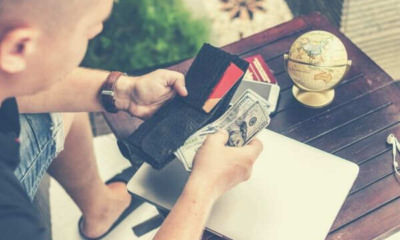 man-holding-u-s-dollar-banknotes-and-black-leather-bi-fold-wallet-laptop-globe-wooden-table