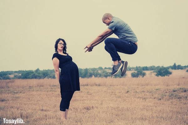 man jumping pointing on womans baby bump grass sky