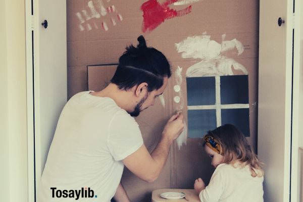 man in white shirt and brown pants painting cardboard house with daughter