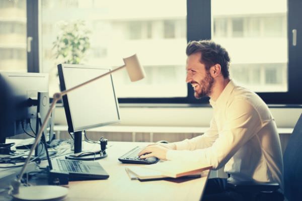 man-in-white-dress-shirt-sitting-office-on-black-rolling-chair-while-facing-black-computer-set-and-smiling