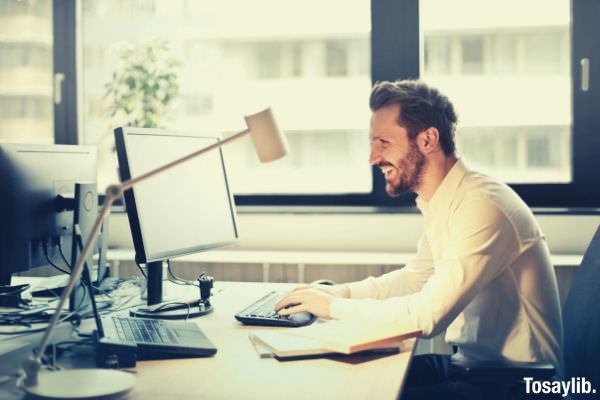 man in white dress shirt sitting on black rolling chair while facing black computer set and smiling