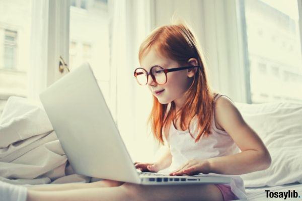 young girl with glasses sitting in bed while sitting laptop