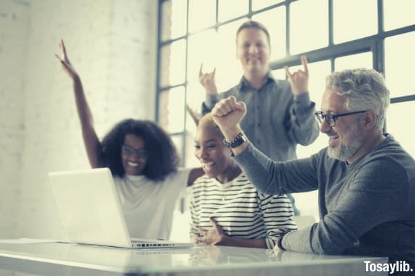 cheerful multiethnic friends with laptop raising hands in success