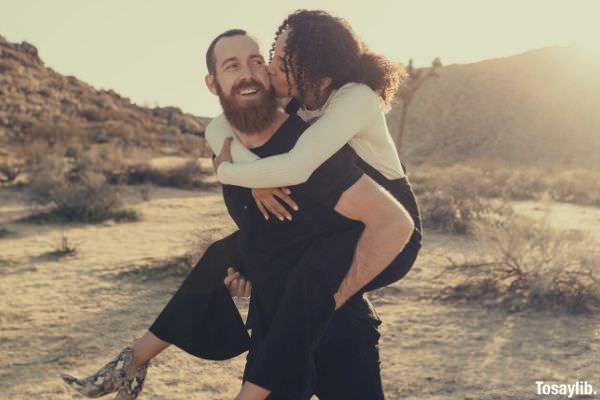 woman in white long sleeves and black pants kissing while on the back of the man in black shirt