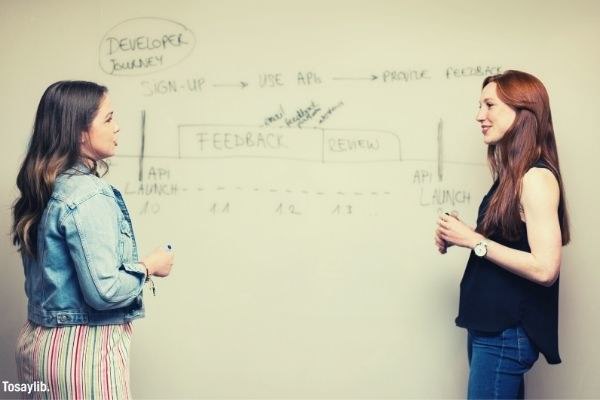 woman in denim jacket and another woman in black sleeveless shirt talking in front of white board