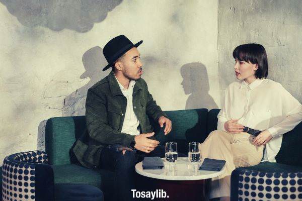 man in green blazer sitting on green sofa while talking to a woman wearing white