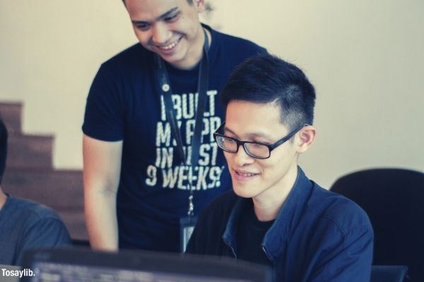 man in black shirt and ID lace standing behind the man sitting with eyeglasses