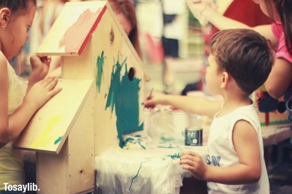 boy in white tank top holding blue green paint painting a bird house with another kids