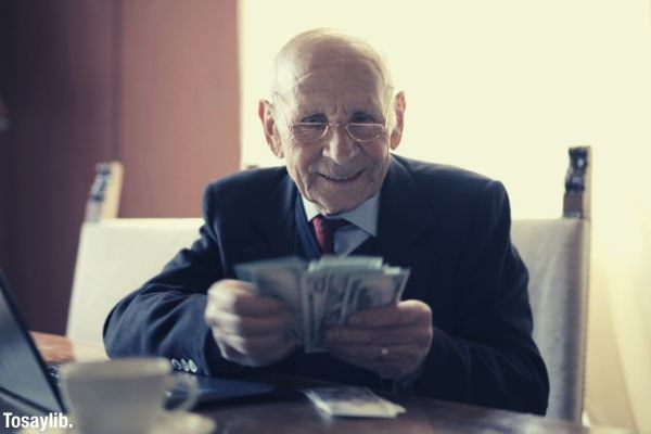 happy senior businessman wearing eyeglasses counting money while sitting at table with laptop