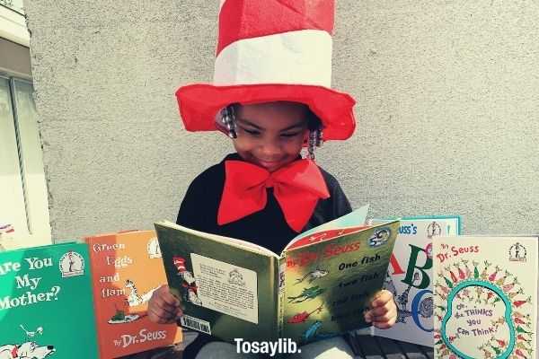 girl in black shirt wearing red and white hat holding Dr Seuss book while sitting on the table