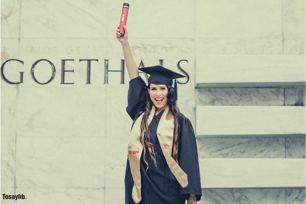 photo of smiling woman in black academic dress standing in front of marble wall holding up diploma