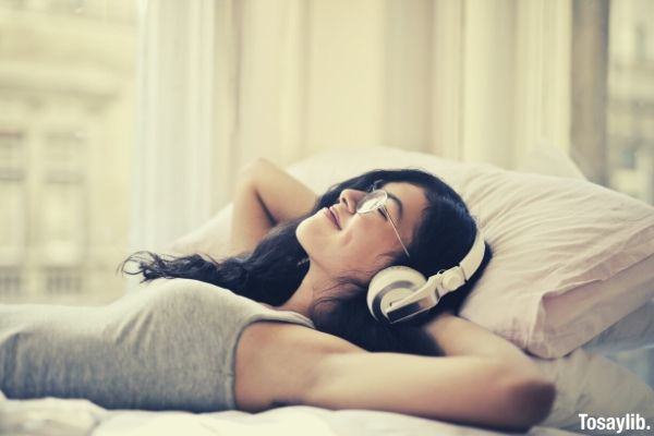 woman in tank top with eyeglasses lying on bed listening to music