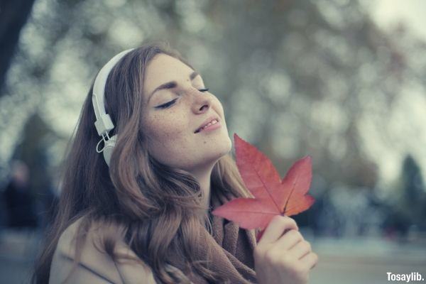 young satisfied woman in headphones with fresh red leaf listening to music with pleasure while lounging in autumn park