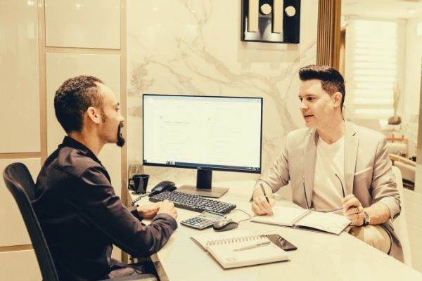 shallow-focus-photo-of-man-in-gray-suit-jacket-taking-notes-talking-to-man-in-black-polo-desktop-white-table