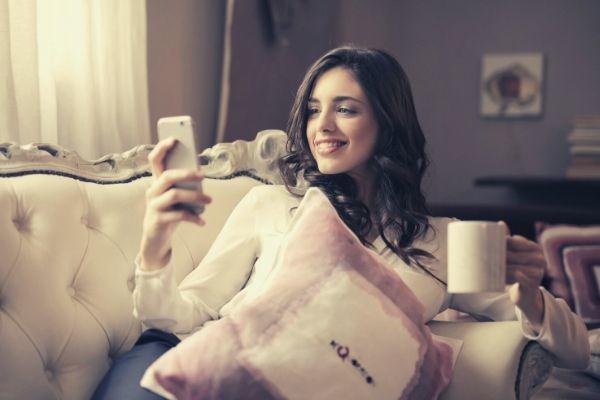 woman-in-white-long-sleeved-shirt-holding-smartphone-and-a-mug-sitting-on-tufted-sofa