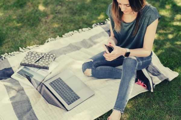 young-student-using-mobile-while-sitting-in-park-with-laptop-picnic-blanket