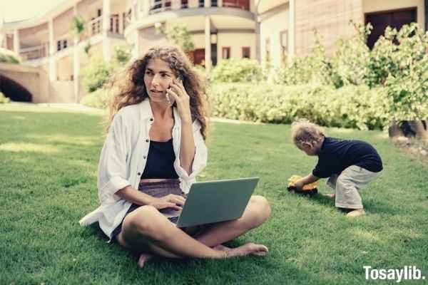 photo of woman using silver laptop with her child playing on grass field talking about something urgent over the phone