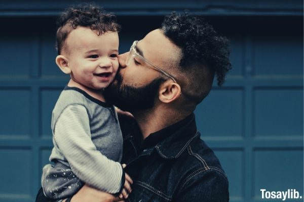 man in black denim polo with undercut hair carrying baby and kissing him