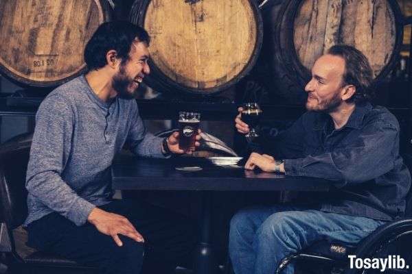 two men sitting beside barrels drinking beer while talking with each other