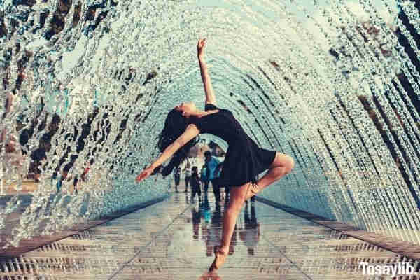 woman in black flare dress dancing raising her hand and foot under water fountain