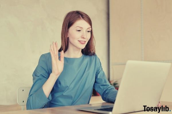 woman in blue long sleeve blouse sitting waving hello on laptop