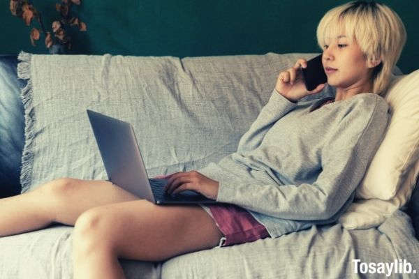 ethnic freelancer typing on laptop and speaking on smartphone wearing longsleeves and shorts sitting on the sofa