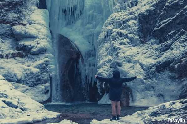 person looking at ice frozen waterfall while spreading arms wide