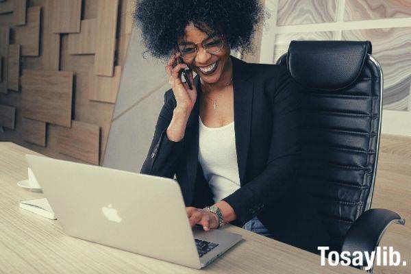 woman in black blazer sitting on black office chair using smart phone smiling