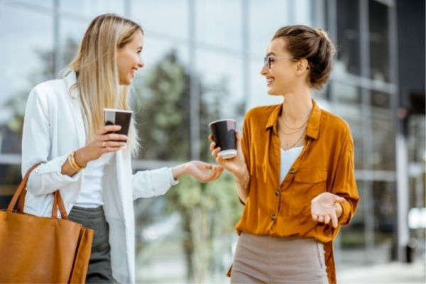 two young businesswomen talking near office while walking holding coffee