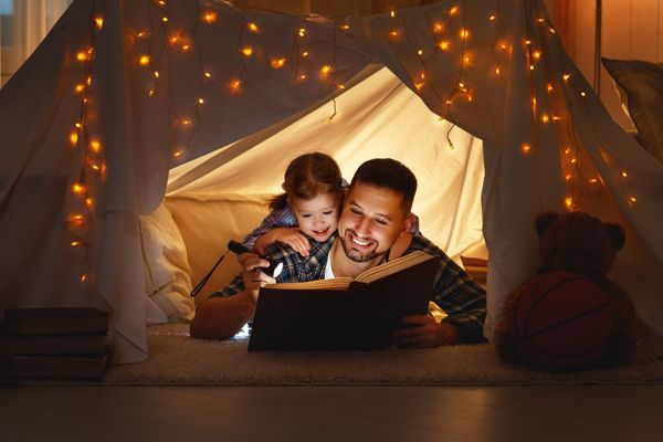 father daughter reading book with flashlight in a tent